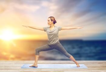 fitness, sport, people and healthy lifestyle concept - woman making yoga warrior pose on pier over sea background. woman making yoga warrior pose on mat