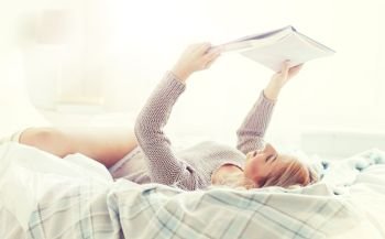 rest, comfort, leisure and people concept - happy young woman reading book in bed at home bedroom. young woman reading book in bed at home