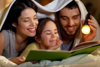 people and family concept - close up of happy mother, father and little daughter reading book with torch light in bed at night at home under blanket. happy family reading book in bed at night at home