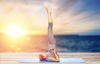 yoga, fitness and people concept - woman making shoulderstand pose on mat outdoors over sea background. woman doing yoga in shoulderstand pose on mat