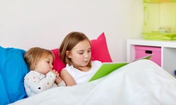 childhood, leisure and family concept - little girls or sisters reading book in bed. little girls or sisters reading book in bed
