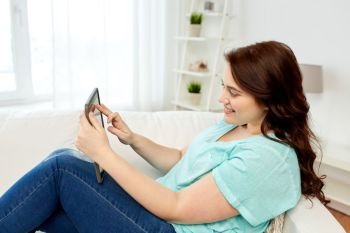 technology, internet and people concept - smiling young plus size woman sitting on couch with tablet pc computer at home. happy young plus size woman with tablet pc at home