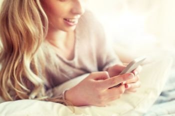 technology, communication and people concept - close up of happy young woman lying in bed and texting on smartphone at home bedroom. close up of woman with smartphone in bed at home