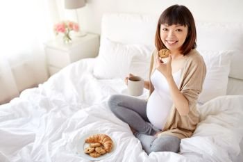 pregnancy, rest, people and breakfast concept - happy pregnant asian woman with cup drinking tea and eating cookie in bed at home bedroom. happy pregnant woman eating cookie in bed at home