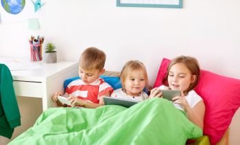 childhood, technology and family concept - little kids with tablet pc computer and smartphones in bed at home. kids with tablet pc and smartphones in bed at home