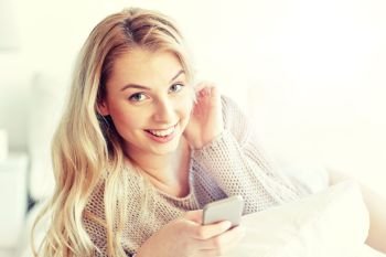 technology, communication and people concept - happy young woman lying in bed with smartphone at home bedroom. happy young woman with smartphone in bed at home