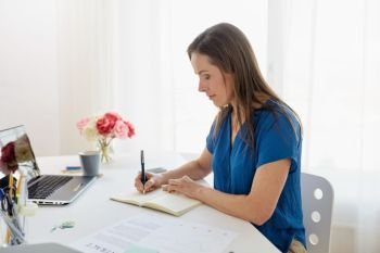 business, people and work concept - middle-aged woman with laptop computer working and writing to notebook at office or home. woman writing to notebook at office