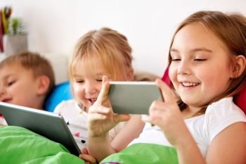 childhood, technology and family concept - happy little kids with tablet pc computer and smartphone in bed at home. happy kids with tablet pc and and smartphone in bed 