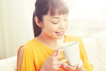 people, drinks and leisure concept - happy asian woman sitting on sofa and drinking tea from cup or mug at home. happy asian woman drinking from tea cup
