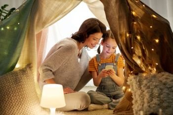 family, hygge and technology concept - happy mother and little daughter with smartphone in kids tent at night at home. happy family with smartphone in kids tent at home