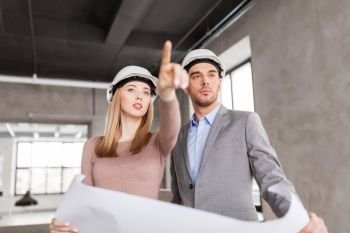 architecture, construction business and people concept - architects with blueprint and helmets at office pointing at something. architects with blueprint and helmets at office