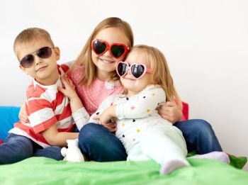 summer, valentines day and childhood concept - happy little kids in heart shaped sunglasses at home. happy little kids in sunglasses at home