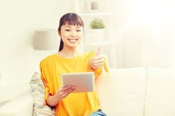 people, technology, gesture and leisure concept - happy young asian woman sitting on sofa with tablet pc computer showing thumbs up at home. happy asian woman with tablet pc and thumbs up. happy asian woman with tablet pc and thumbs up