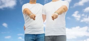 lgbt, same-sex relationships and homosexual concept - close up of hugging male couple wearing gay pride rainbow awareness wristbands and pointing fingers at you over blue sky and clouds background. couple with gay pride rainbow wristbands. couple with gay pride rainbow wristbands