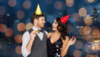 birthday, celebration and holidays concept - happy couple with party blowers and caps having fun over singapore city night lights background. happy couple with party blowers having fun. happy couple with party blowers having fun