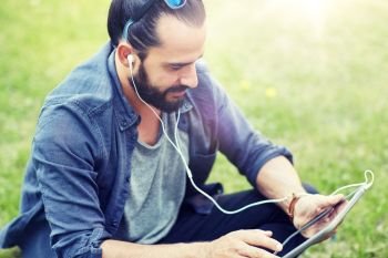 people, music, technology, leisure and lifestyle - happy young hipster man with earphones and tablet pc computer sitting on grass. man with earphones and tablet pc sitting on grass. man with earphones and tablet pc sitting on grass