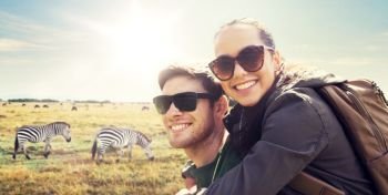 travel, tourism and people concept - happy couple with backpacks having fun over african savannah and zebras background. smiling couple with backpacks traveling in africa. smiling couple with backpacks traveling in africa