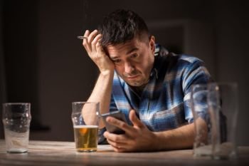 alcoholism, alcohol addiction and people concept - male alcoholic with smartphone drinking beer and smoking cigarette at night. man with cellphone drinking alcohol and smoking. man with cellphone drinking alcohol and smoking