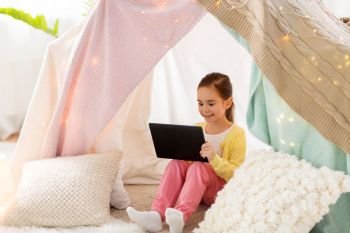 childhood, technology and hygge concept - happy little girl with tablet pc computer in kids tent at home. little girl with tablet pc in kids tent at home. little girl with tablet pc in kids tent at home