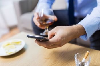 alcoholism, alcohol addiction and technology concept - close up of male hands with smartphone and brandy at home. man with smartphone and glass of alcohol at home. man with smartphone and glass of alcohol at home
