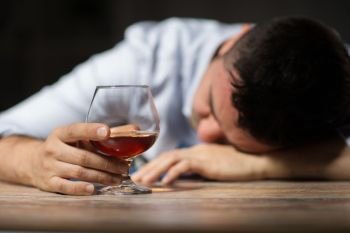 alcoholism, alcohol addiction and people concept - male alcoholic with glass of brandy lying or sleeping on table at night. drunk man with glass of alcohol on table at night. drunk man with glass of alcohol on table at night