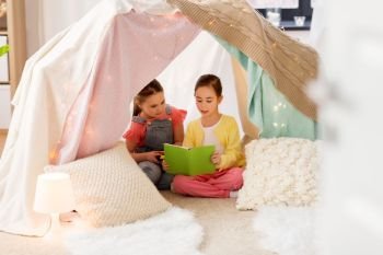 childhood, friendship and hygge concept - happy little girls reading book in kids tent or teepee at home. little girls reading book in kids tent at home. little girls reading book in kids tent at home