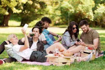 friendship, leisure, technology and people concept - group of friends with smartphones chilling on picnic blanket and drinking non alcoholic beer at summer park. friends with smartphones on picnic at summer park. friends with smartphones on picnic at summer park
