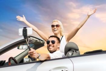 road trip, technology and travel concept - happy couple driving in convertible car and taking picture by smartphone on selfie stick over sky background. couple in car taking selfie by smartphone over sky. couple in car taking selfie by smartphone over sky
