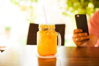 A cup full of fresh orange juice with cocktail straw on wooden table. Man is doing mobile photography. Fresh orange pleasure