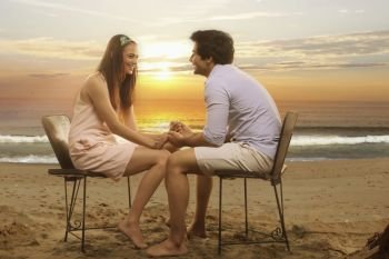 Happy couple sitting in chairs holding hands on beach at sunset