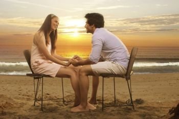 Young couple sitting in chairs holding hands on beach at sunset