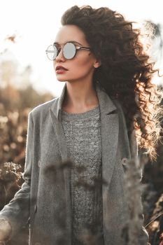 Outdoor fashion photo of young beautiful lady in autumn landscape with dry flowers. Gray coat, knitted sweater, sunglusses, wine lipstick. Fashion lookbook. Warm Autumn. Warm Spring