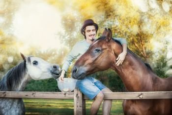 Young happy guy sitting on fence, laughing and feeding and hugging horses  at country nature background