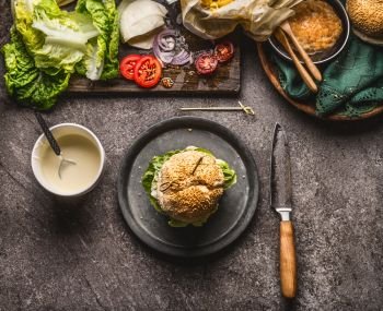 Tasty homemade burger on plate with knife and ingredients on dark rustic background, top view