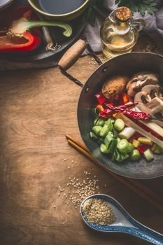 Wok pot with vegetarian asian cuisine ingredients for stir fry with chopped vegetables, spices, sesame seeds and chopsticks on rustic wooden background, top view. Chinese or Thai food cooking concept
