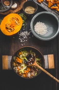 Cooking preparation with  pot of vegetarian pumpkin risotto and spoon on dark rustic kitchen table background with cooking ingredients, top view. Healthy clean seasonal food and eating concept