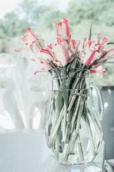 Bunch of tulips in glass jug on white table at window. Flowers in interior design. Cozy home. Springtime
