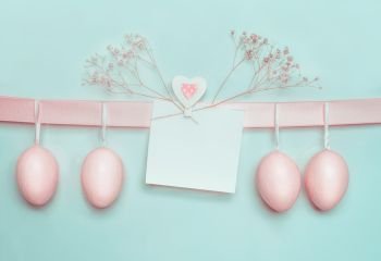Easter greeting card mock up with pastel pink eggs hanging on ribbon on light  blue turquoise background. Creative greeting concept. Layout with copy space for your text