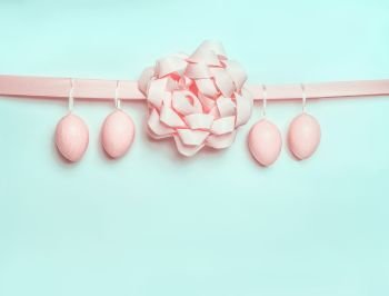 Pastel pink Easter eggs hanging on ribbon with beautiful bow on light  blue turquoise background. Creative greeting concept. Layout with copy space for your text