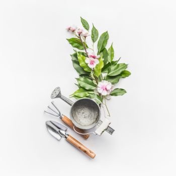Watering can with gardening tools and green bunch of twigs with blossom decoration on white desk background, top view