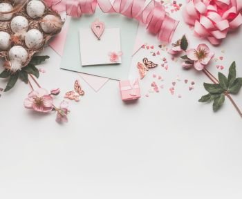 Modern styled Easter background with pastel color decoration: eggs, ribbon, flowers, bow and gift box. Mock up for Easter greeting card on white desk background, top view