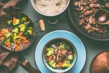 Healthy balanced nutrition dish in pots and bowls with beef meat, steamed vegetables and rice on table background with plate and cutlery, top view