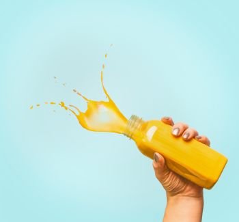 Female hand holding bottle with yellow splash summer beverage: smoothie or juice at blue background, copy space