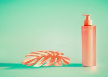 Pink cosmetic bottle with  dispenser pump and tropical leaf at mint background. Summer skin care or sunblock product with copy space for design
