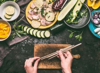 Chicken meat and vegetables skewers making. Female hands holding wooden skewer on kitchen table background with chicken pieces and vegetables, top view. Grill preparation