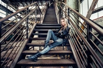 Beautiful woman posing wearing casual outfit with leather jacket, black shoes and fashionable jeans. Girl posing in industrial plant. 