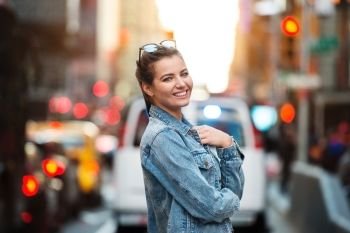 Lifestyle photo of cute happy smiling woman on standing on city street. Woman wearing jeans jacket and sunglasses outdoors at sunset time.
