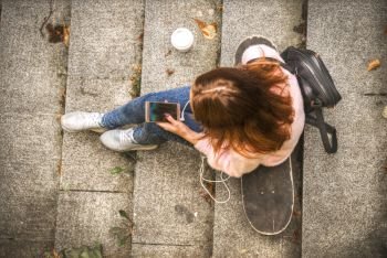 girl is sitting on a skateboard in headphones with a glass of coffee and a smartphone. View from above. girl is sitting on a skateboard in headphones with a glass of co