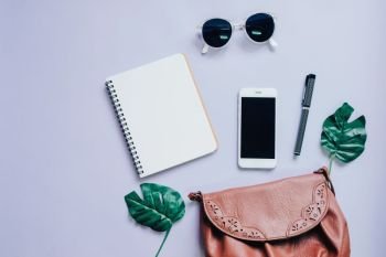 Top view flat lay of woman bag with smartphone, blank notebook, sunglasses and green leaf, copy space