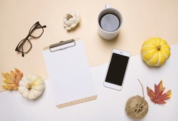 Creative flat lay of workspace desk in autumn style with blank clipboard, smartphone, eyeglasses, pumpkins, coffee and autumn leaves with copy space background, minimal style